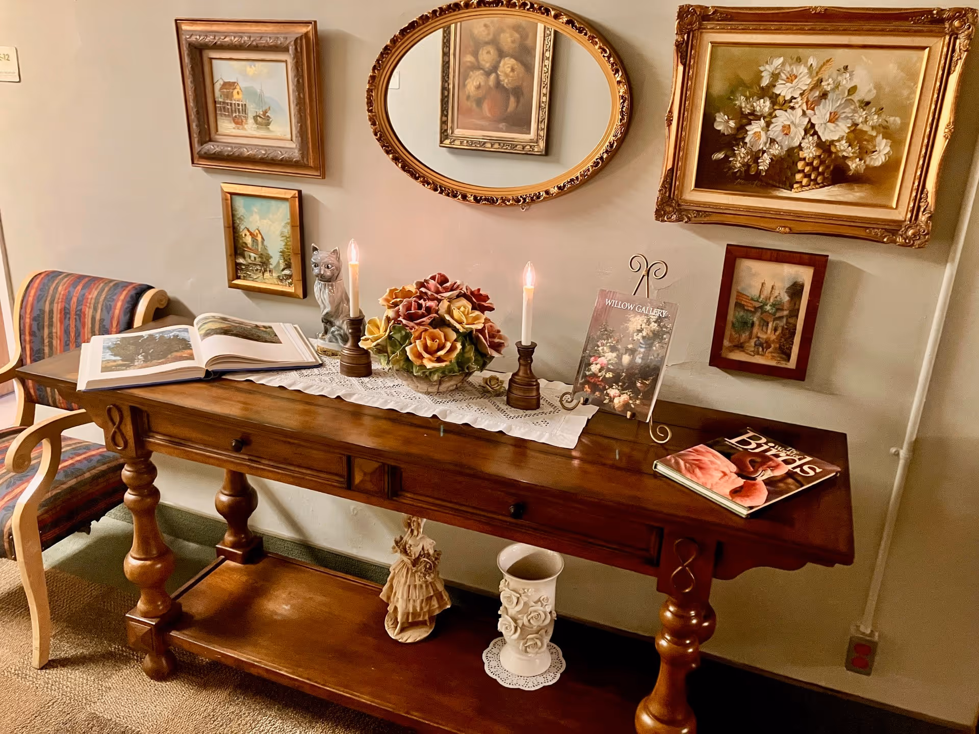 A wooden console table with turned legs and three drawers, decorated with a lace runner, a ceramic flower arrangement, two lit candles in holders, an open book, a closed book titled 'Birds', and a small framed picture on a stand. Above the table are five framed paintings, including an oval mirror reflecting a floral painting. To the left of the table is a wooden chair with striped upholstery.