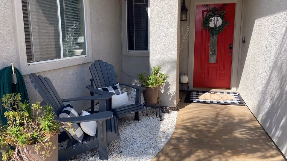 Outdoor entrance area of a residential care home featuring a red front door decorated with a wreath, two black Adirondack chairs with black and white checkered pillows, potted plants, white gravel ground cover, and a garden hose coiled on the wall.