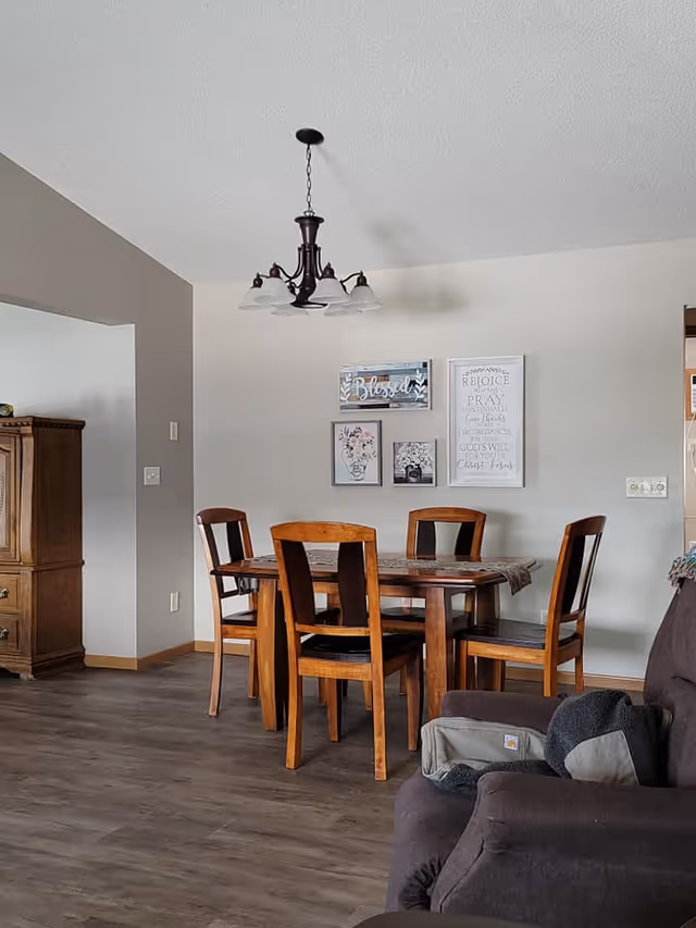 A dining area with a wooden table and four matching wooden chairs with black cushions. Above the table hangs a dark metal chandelier with five lights. On the wall behind the table are four framed decorative art pieces with inspirational and floral themes. To the left is a wooden cabinet, and to the right is part of a dark upholstered armchair with a gray pillow and a small bag on it. The floor is a wood-like laminate, and the walls are painted light gray.
