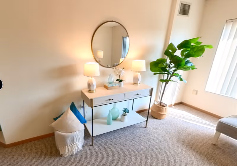 A bright interior corner of a room featuring a console table with two lamps, decorative vases, and a small plant. Above the table is a round mirror reflecting part of the room. To the right, there is a large potted plant near a window with vertical blinds, and a cushioned chair is partially visible.