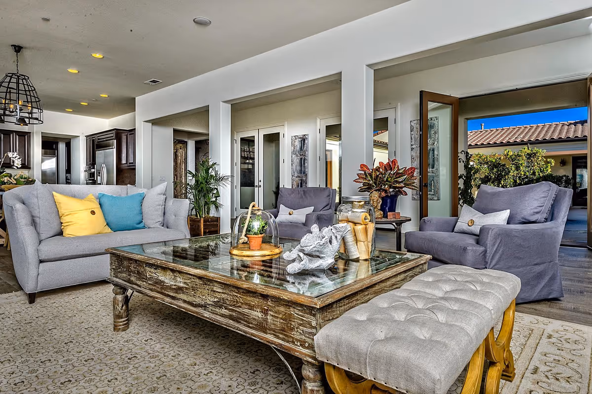 A bright and spacious living room featuring a gray sofa with yellow and blue pillows, two gray armchairs, a rustic wooden coffee table with decorative items, and a cushioned bench. The room opens to an outdoor patio with greenery and a tiled roof visible through glass doors. The kitchen area with dark wood cabinets and stainless steel appliances is visible in the background.