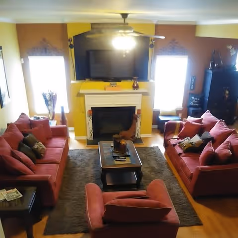 Cozy living room with red sofas arranged around a coffee table facing a fireplace and wall-mounted TV, with windows on either side.