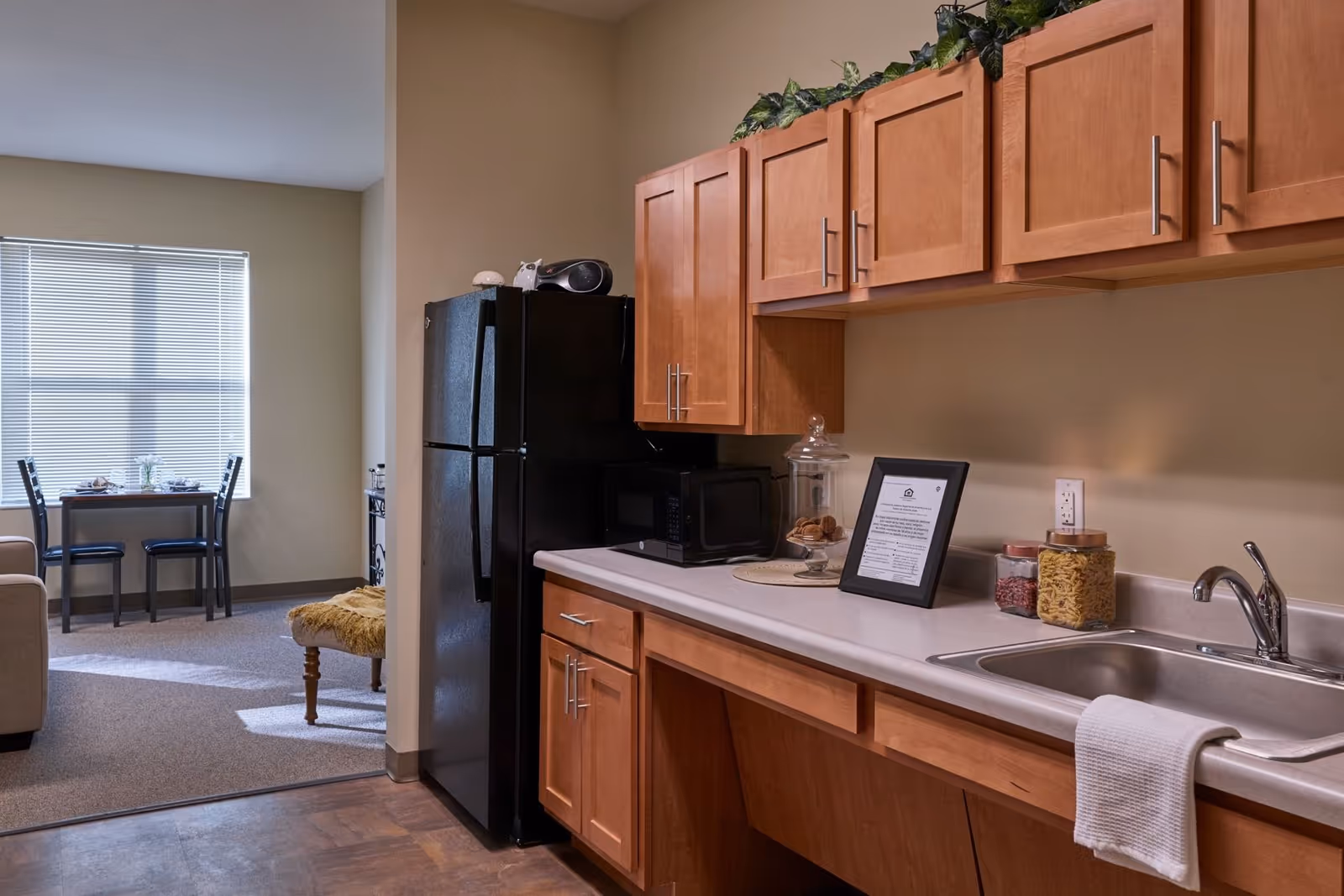 A kitchen area with wooden cabinets, a black refrigerator, a microwave, a sink with a white towel hanging on it, and jars containing pasta and beans on the countertop. In the background, there is a dining area with a small table and two chairs near a window with blinds.
