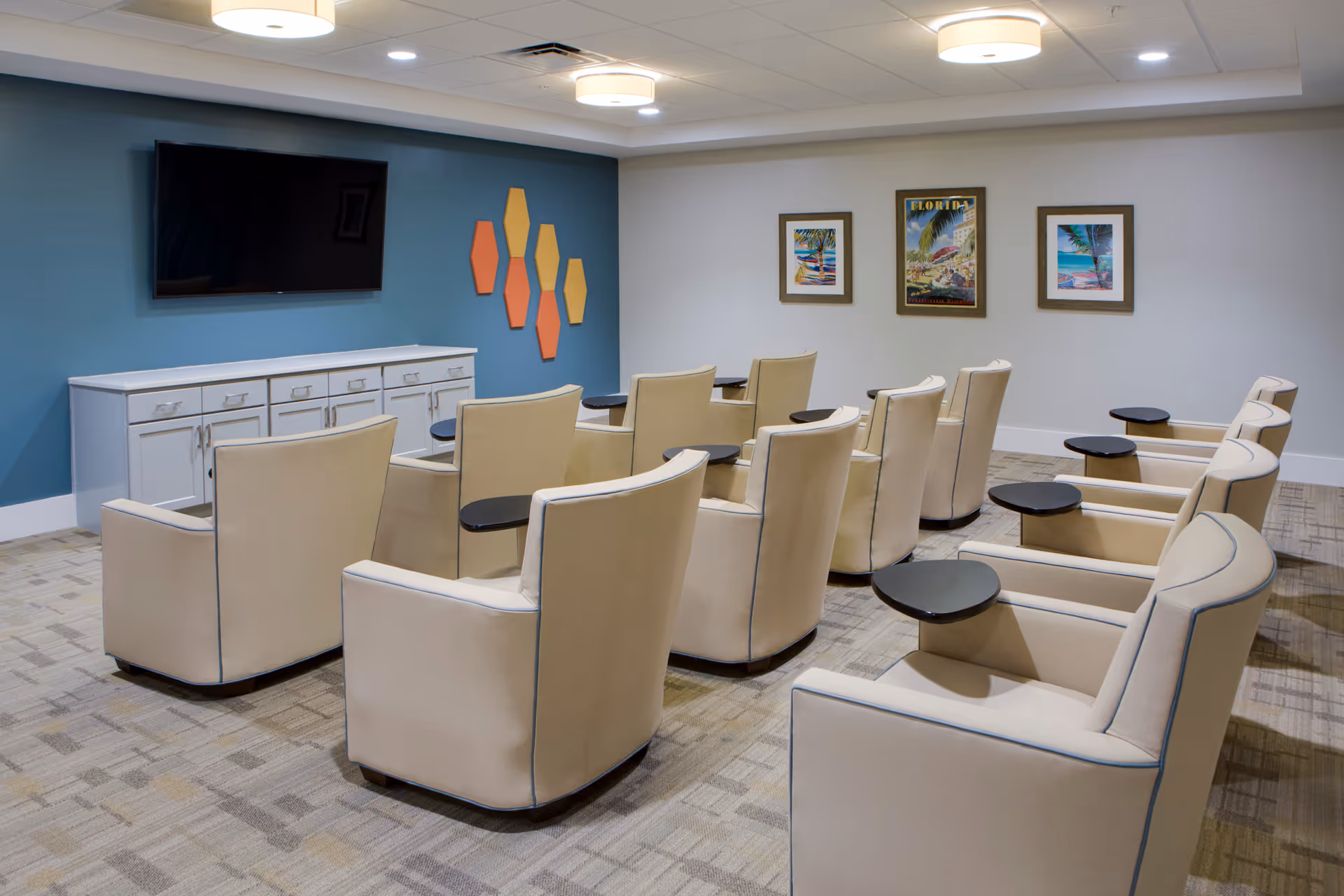A small theater or media room with two rows of beige armchairs, each equipped with a small black side table. A large flat-screen TV is mounted on a blue accent wall above a white cabinet. The opposite wall is white and decorated with three framed tropical-themed pictures. The room has a patterned carpet and ceiling lights.