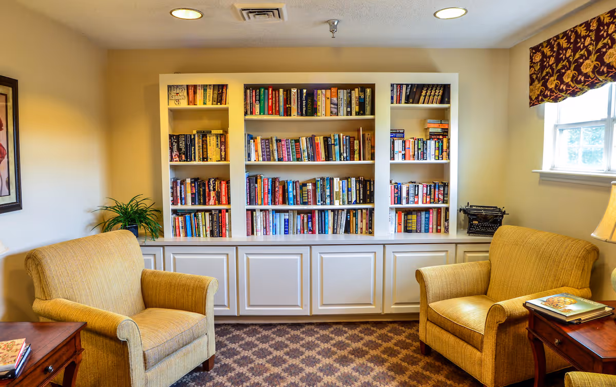A cozy reading nook in a senior living community featuring two beige upholstered armchairs facing each other with wooden side tables beside them. Behind the chairs is a built-in white bookshelf filled with books. A window with a patterned valance allows natural light into the room, and a table lamp is visible on the right side. The room has a patterned carpet and soft yellow walls.
