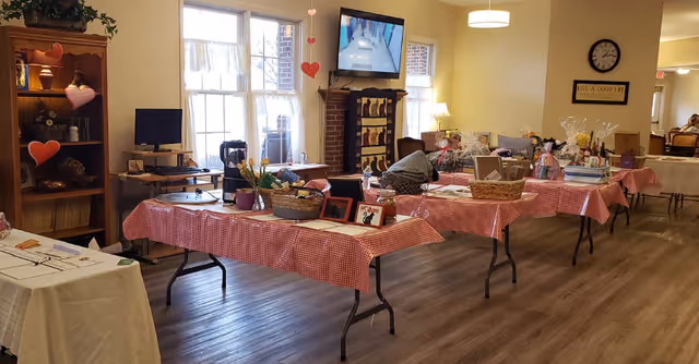 Interior room with several tables covered in red and white checkered tablecloths displaying various items such as baskets, framed pictures, and decorations. A wooden shelf with plants and decor is on the left, and a TV mounted above a brick fireplace is visible in the background. The room has large windows letting in natural light and a wall clock with a sign that reads 'LIVE A GOOD LIFE'.