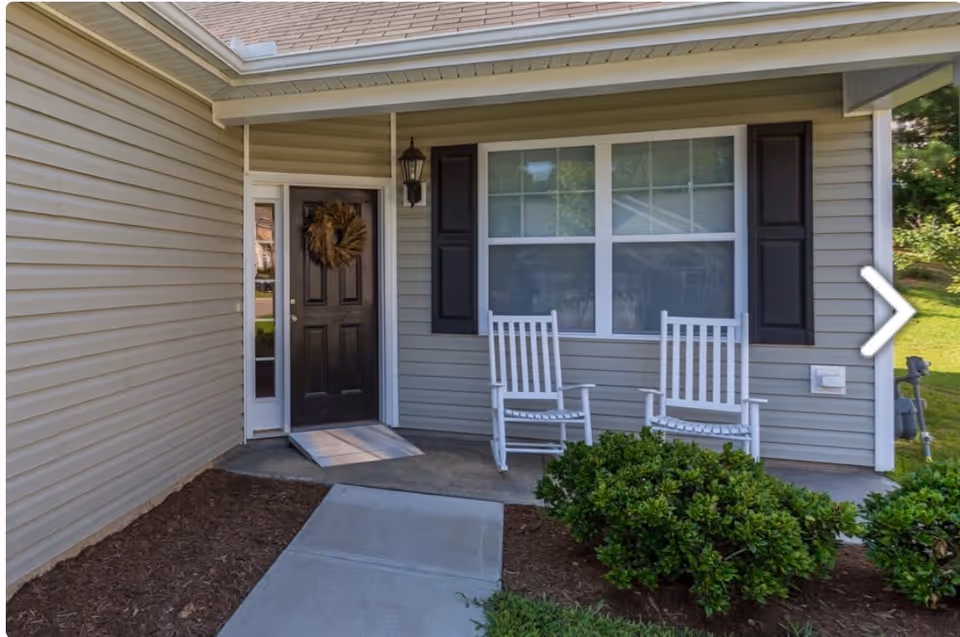 Front porch of a house with a black door decorated with a wreath, two white rocking chairs, a large window with black shutters, a hanging lantern light, and green bushes near the concrete walkway.