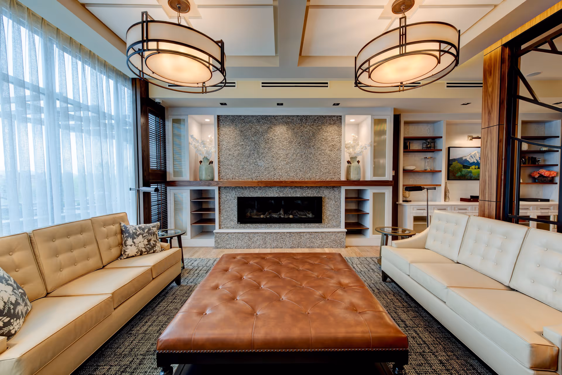 A modern living room with two beige sofas facing each other, a large brown tufted leather ottoman in the center, and a stone fireplace with built-in shelves on either side. The room has large windows with sheer curtains, two round ceiling lights, and decorative vases on the shelves.
