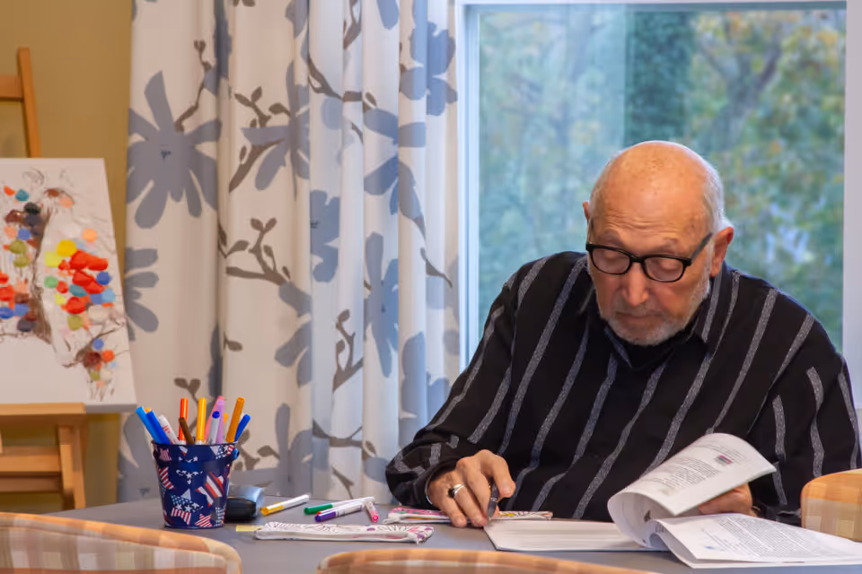 An elderly man wearing glasses and a black striped shirt is sitting at a table, reading and coloring in a book with colored markers. Behind him is a window with a view of trees and curtains with a floral pattern. On the table are various colored markers and a small container holding more markers. An easel with a colorful butterfly artwork is visible in the background.