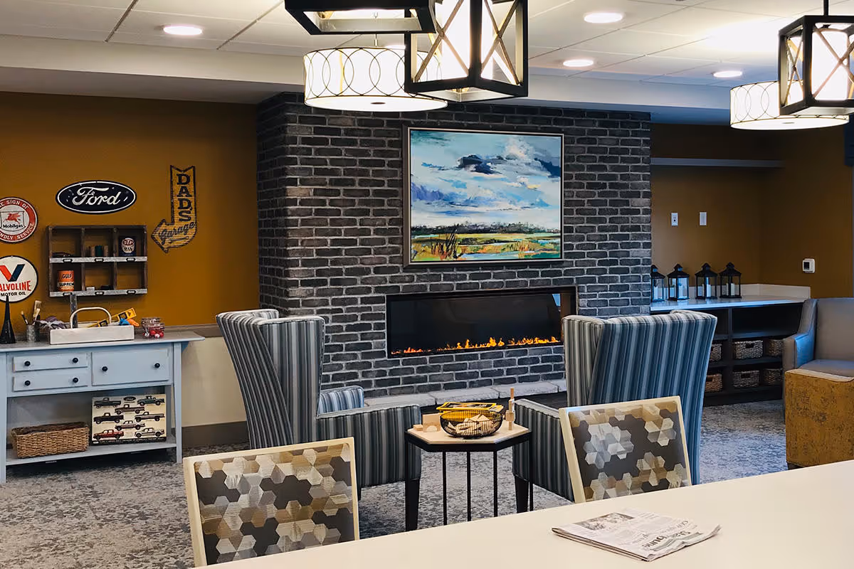 A cozy interior lounge area with a modern electric fireplace set in a dark brick wall. Above the fireplace is a colorful landscape painting. Two striped armchairs face the fireplace with a small table between them holding a decorative basket. The room has warm yellow walls with vintage automotive signs and a white sideboard with shelves and baskets. Ceiling lights with geometric shades illuminate the space.