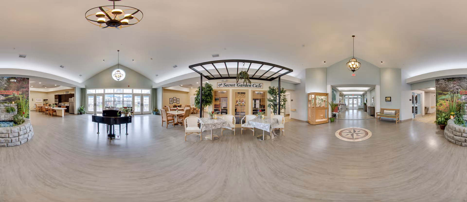 A spacious and well-lit senior living facility common area featuring a black grand piano in the center, several tables and chairs arranged for dining or socializing, and a café area labeled 'Secret Garden Cafe'. The room has large windows allowing natural light, decorative plants, and a compass design on the floor near a hallway entrance.