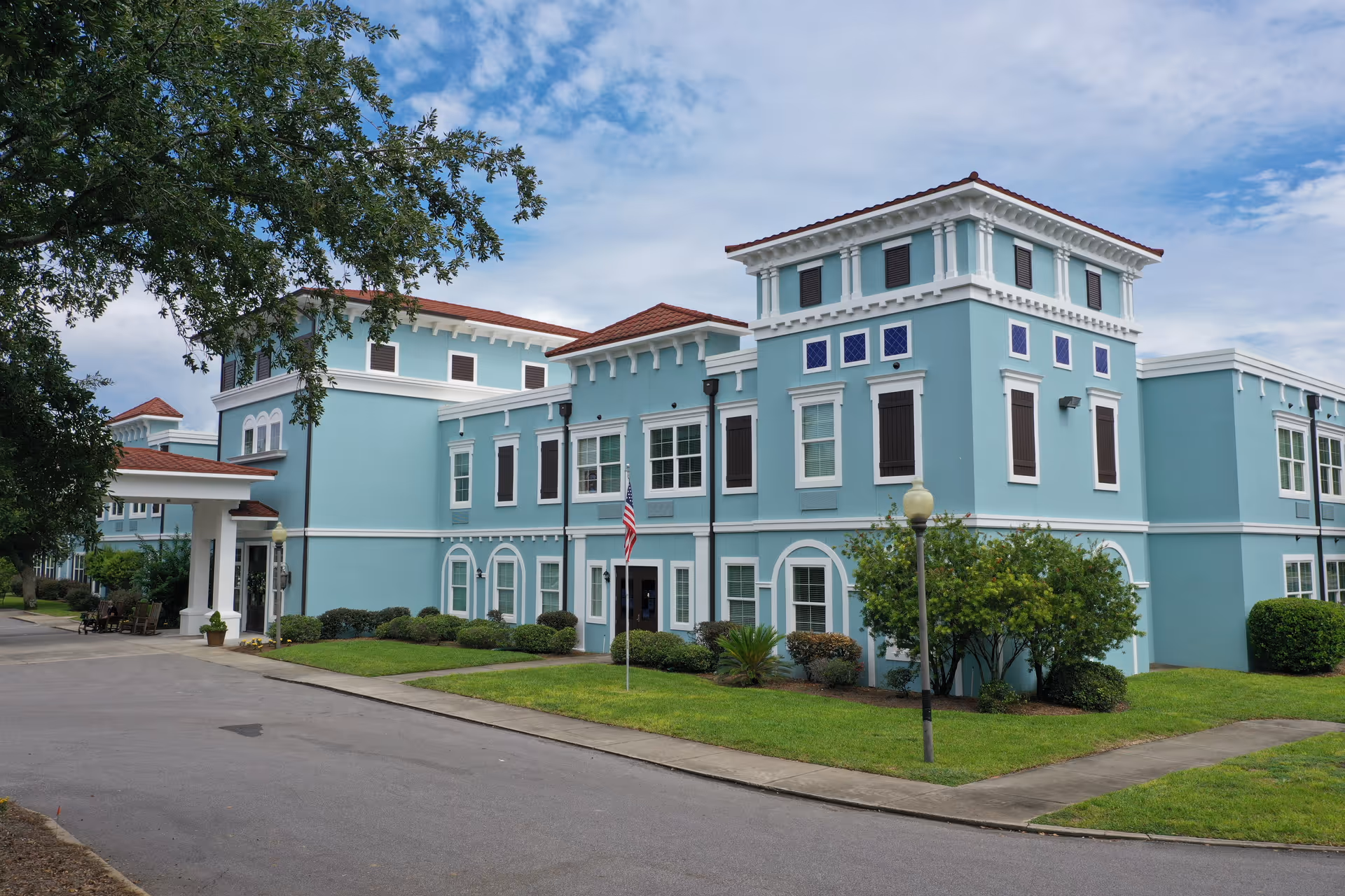 Blue multi-story senior living facility exterior with manicured lawn, shrubs, and an American flag.