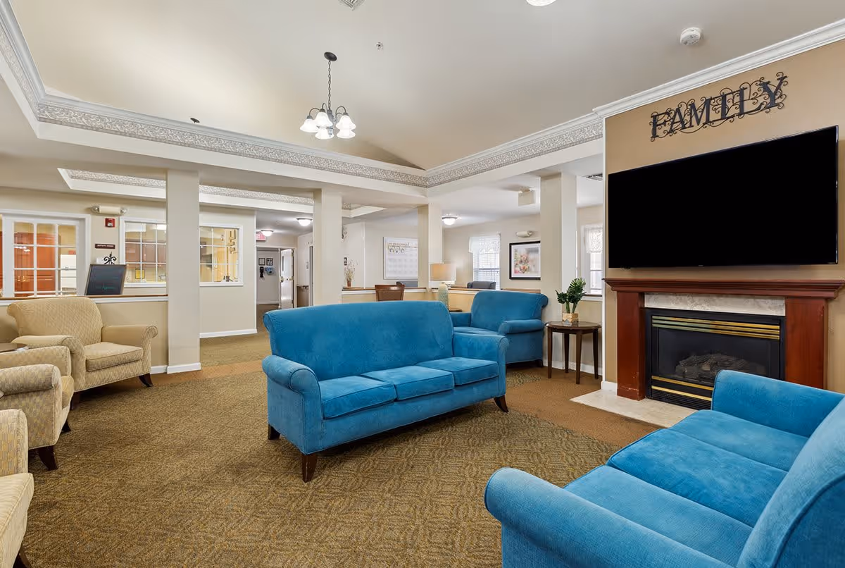 A spacious living room area in a senior living facility with blue upholstered sofas and beige armchairs arranged around a fireplace with a large flat-screen TV mounted above it. The word 'FAMILY' is displayed on the wall above the TV. The room has beige walls, carpeted floors, and ceiling lights, with windows allowing natural light to enter.