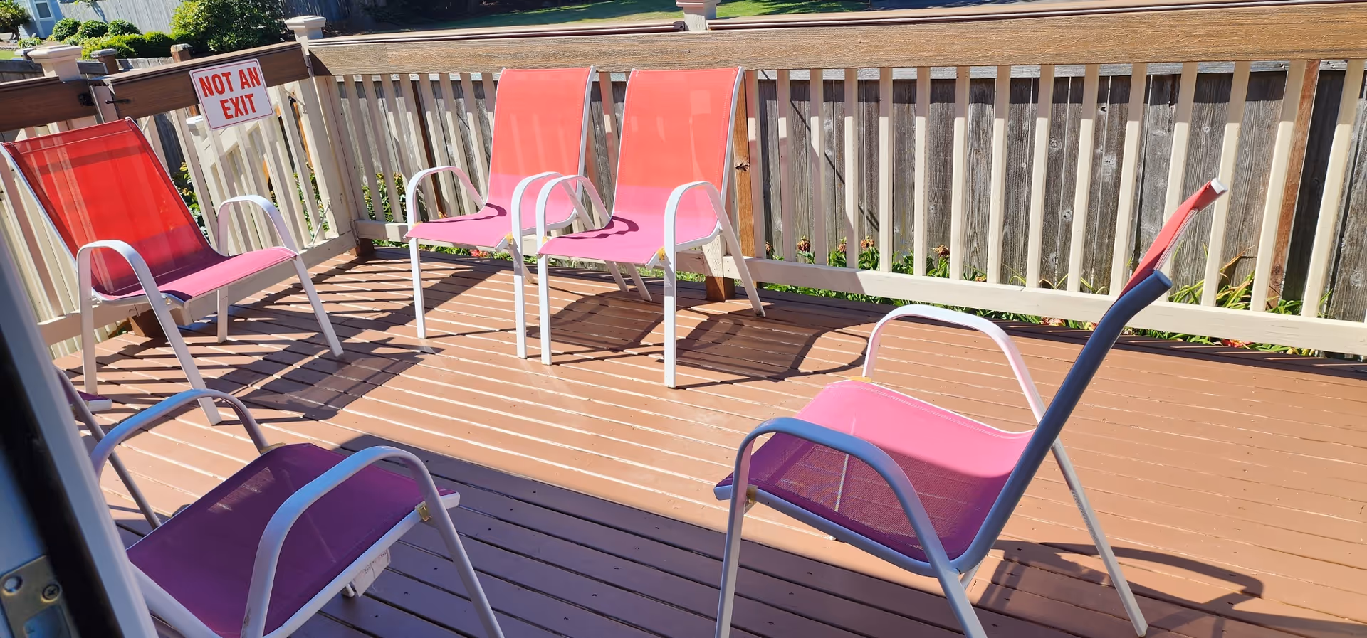 Outdoor wooden deck area with five pink and red mesh chairs arranged around the space. A wooden railing surrounds the deck, and a sign on the railing reads 'NOT AN EXIT'. There is a wooden fence and some greenery visible beyond the deck.