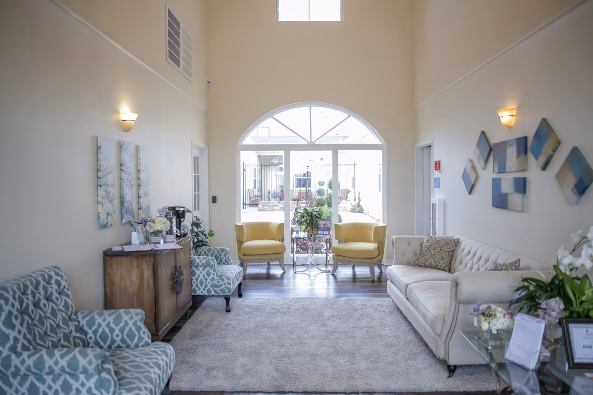 A bright and inviting living room area with a high ceiling, featuring two yellow armchairs and a small glass table in front of a large arched window. There is a beige sofa with patterned cushions on the right, two blue patterned armchairs on the left, a wooden cabinet with a coffee maker and flowers, and abstract wall art on both sides. The floor has a light-colored rug and wood flooring.