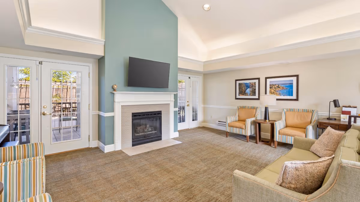 A bright and spacious living room with a high ceiling, featuring a green accent wall with a mounted flat-screen TV above a white fireplace. The room has beige carpeting, a green sofa with pillows, two striped armchairs, and two tan armchairs around a small wooden side table with a lamp. French doors lead to an outdoor patio area with a table and chairs. Two framed landscape pictures hang on the far wall.