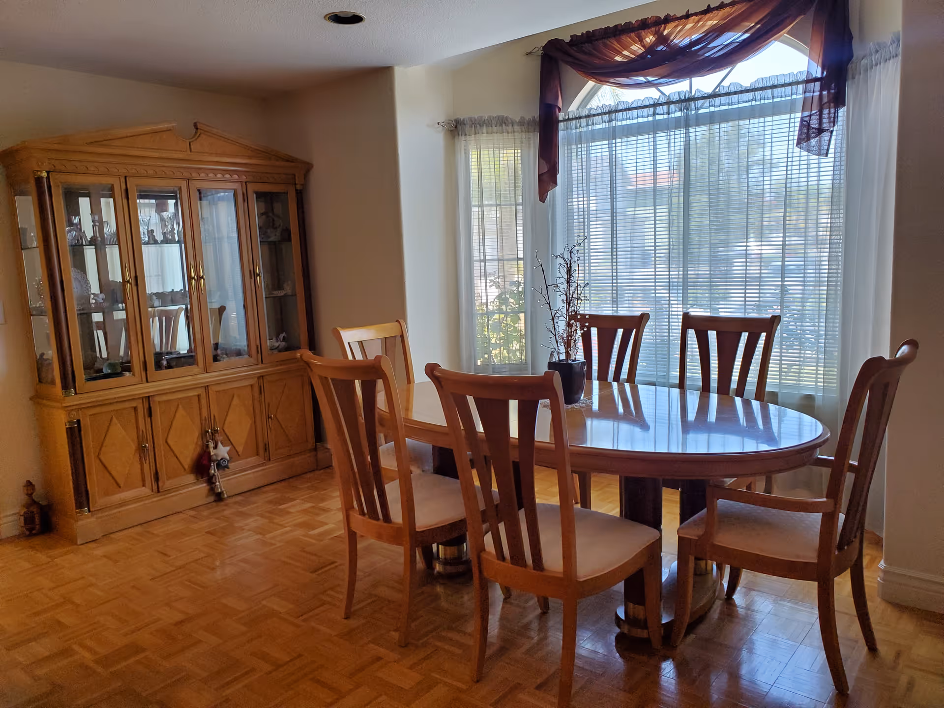 A dining room with a wooden oval dining table surrounded by six wooden chairs with cushioned seats. Behind the table is a large window with blinds and sheer curtains, topped with a decorative valance. To the left of the table is a wooden china cabinet displaying glassware and decorative items. The floor is parquet wood.