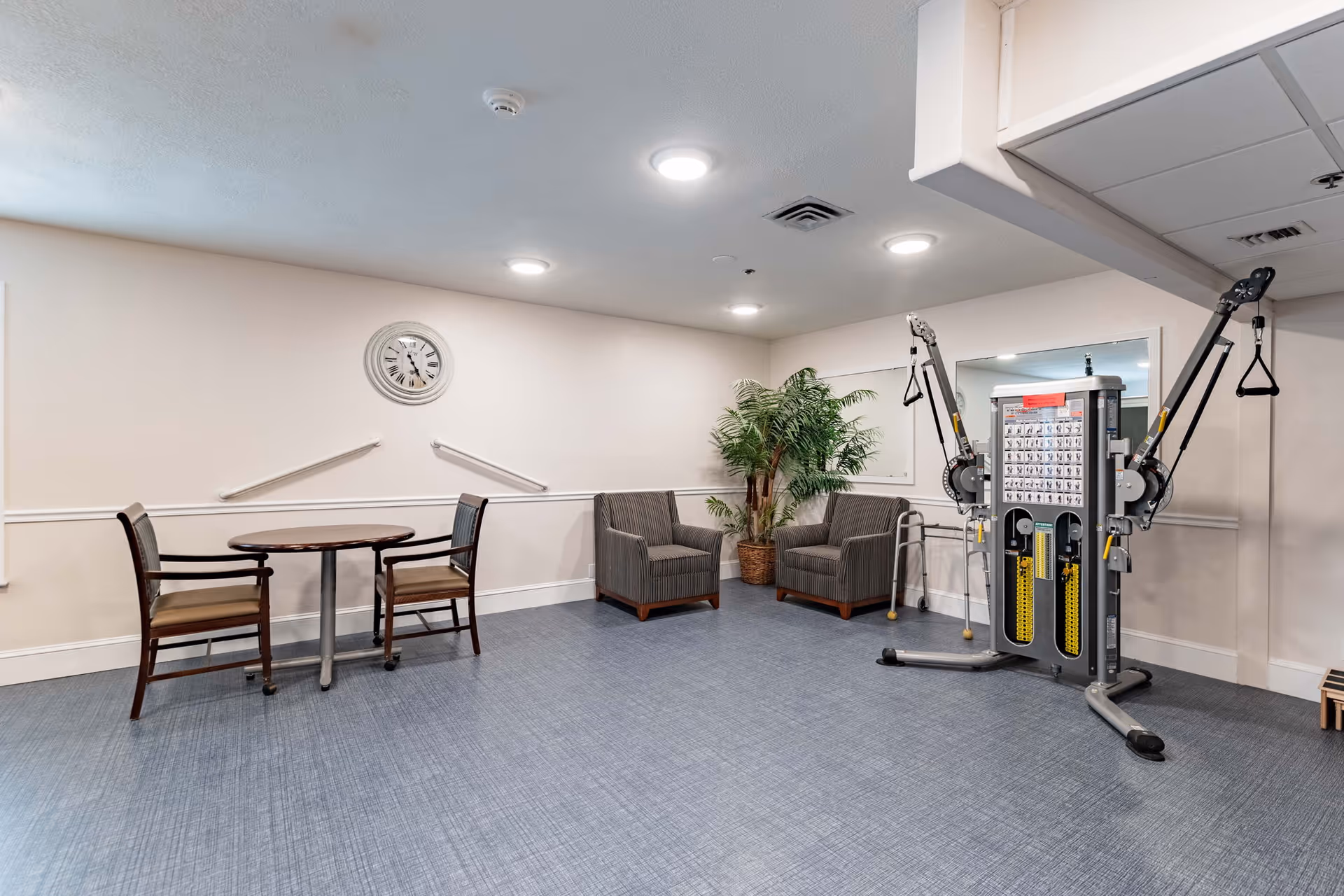 Interior common room with a small round table and chairs, two armchairs by a potted plant, and an exercise machine against the wall.