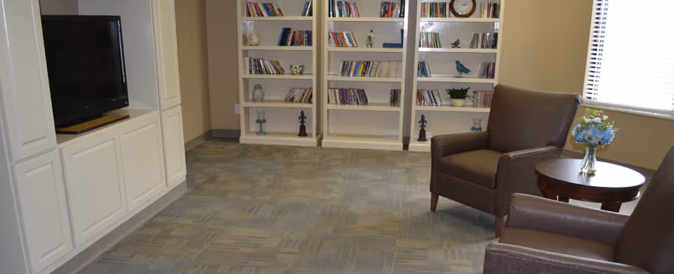 A cozy living room area with two brown armchairs and a round wooden side table with a vase of blue flowers. There is a built-in white entertainment center with a flat-screen TV on the left and white bookshelves filled with books and decorative items along the back wall. A window with blinds allows natural light into the room.