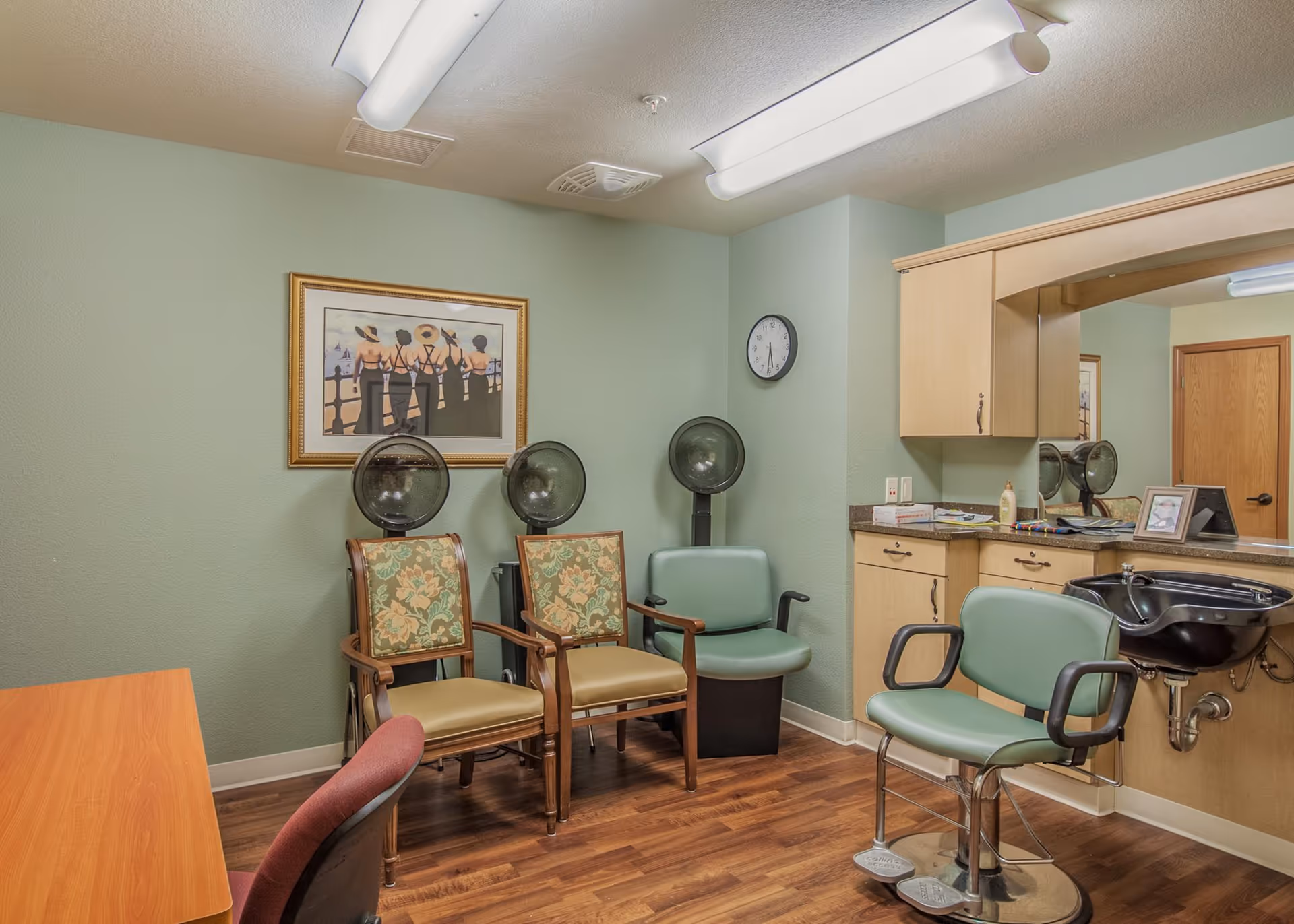 Interior of a hair salon area in a senior living facility with three vintage hair dryers, two floral upholstered chairs, a green salon chair, a black shampoo sink, a large mirror, wooden cabinets, and a framed painting of women in black dresses and hats on the wall.
