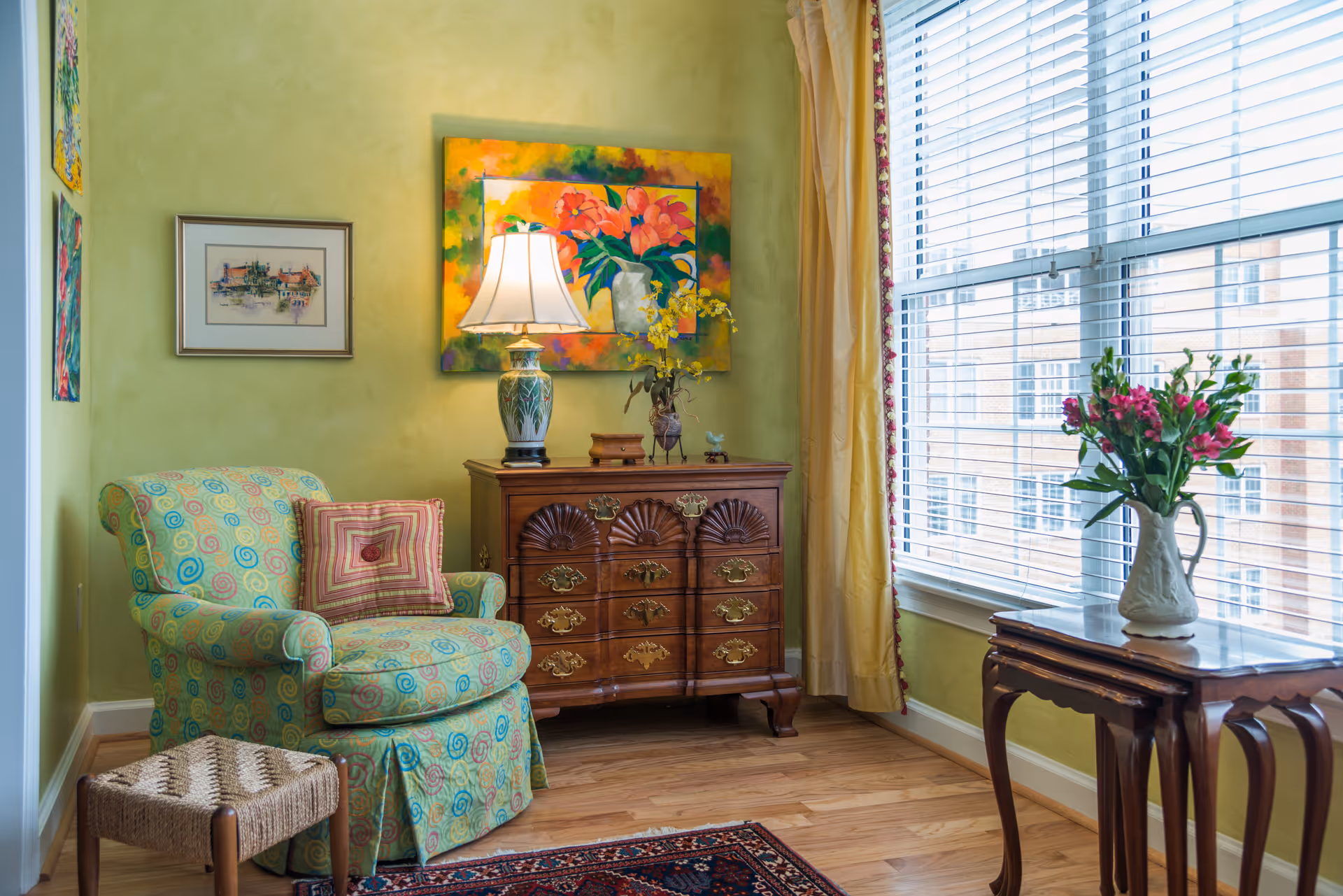 Sunlit living room corner with a patterned armchair, wooden chest topped by a lamp and colorful painting, and a vase of flowers on a side table by the window.