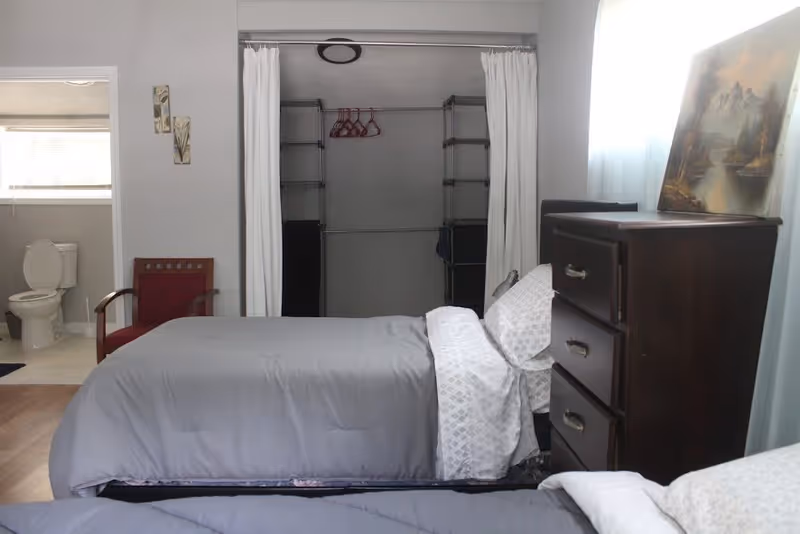 Twin bedroom with gray bedding, a dark wooden dresser, an open closet and an adjacent bathroom visible.