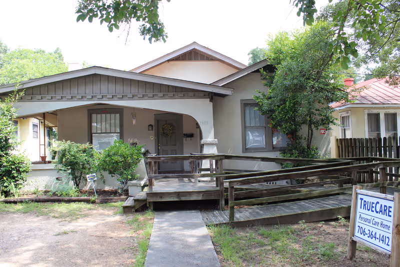 Front exterior view of a single-story personal care home with a covered porch and a wooden ramp leading to the entrance. The building is surrounded by trees and bushes, and there is a sign in the front yard that reads 'TrueCare Personal Care Home' with a phone number.