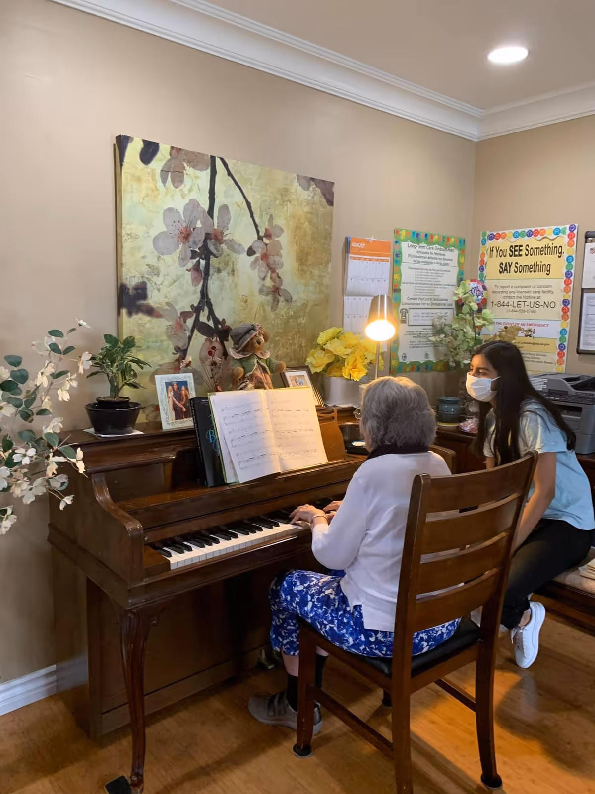 An elderly woman playing a wooden piano with sheet music in front of her, while a younger woman wearing a face mask sits beside her. The room has beige walls decorated with a large floral painting, a calendar, informational posters, and various plants and framed photos on the piano.