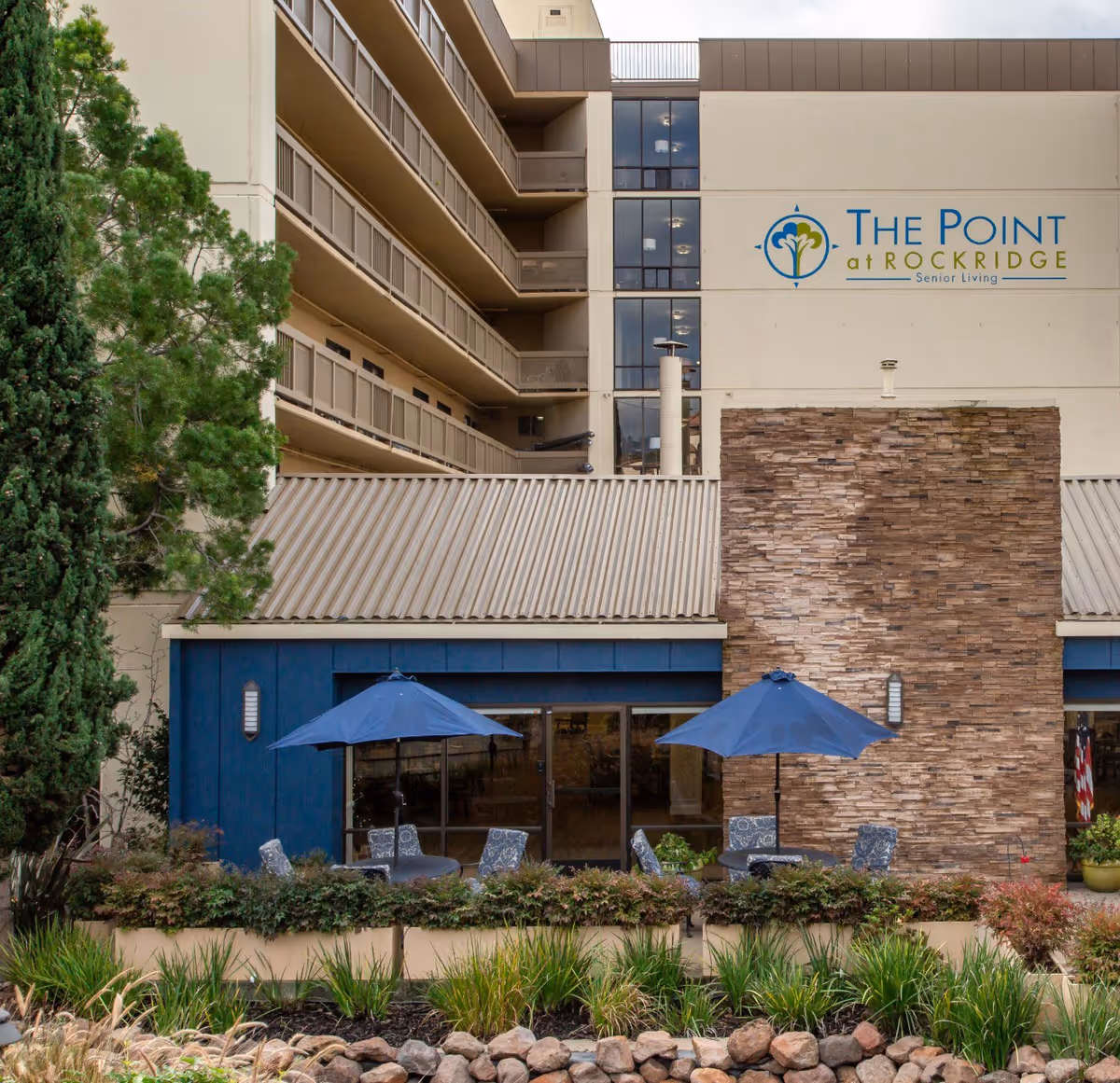 Outdoor patio area at The Point At Rockridge Senior Living featuring two tables with blue umbrellas and chairs, surrounded by greenery and plants. The building facade includes a stone wall section and multiple balconies above.