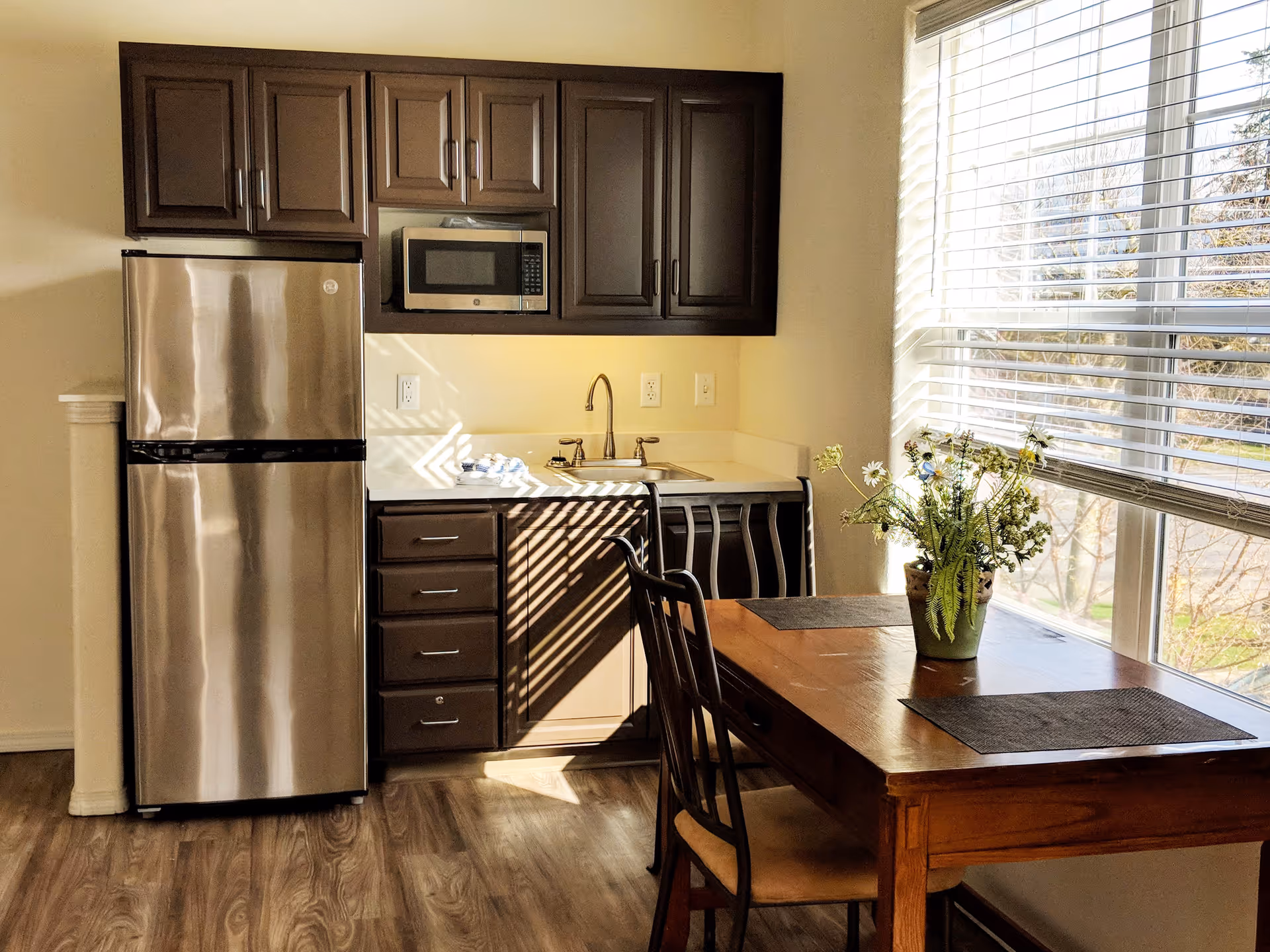 Small kitchenette with stainless steel refrigerator, microwave, sink, dark cabinets and a wooden dining table by a large window with blinds and a vase of flowers.