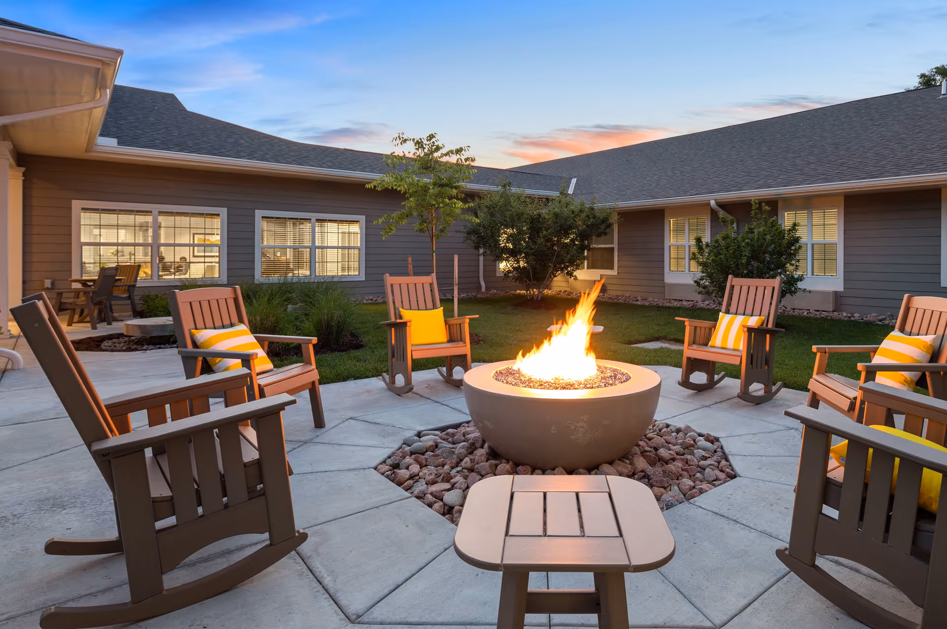 Outdoor patio area at sunset with a circular fire pit surrounded by six wooden rocking chairs, each with a yellow and white striped cushion. The patio is part of a single-story building with gray siding and multiple windows, some with lights on inside. There are small trees and bushes in the grassy area around the patio.
