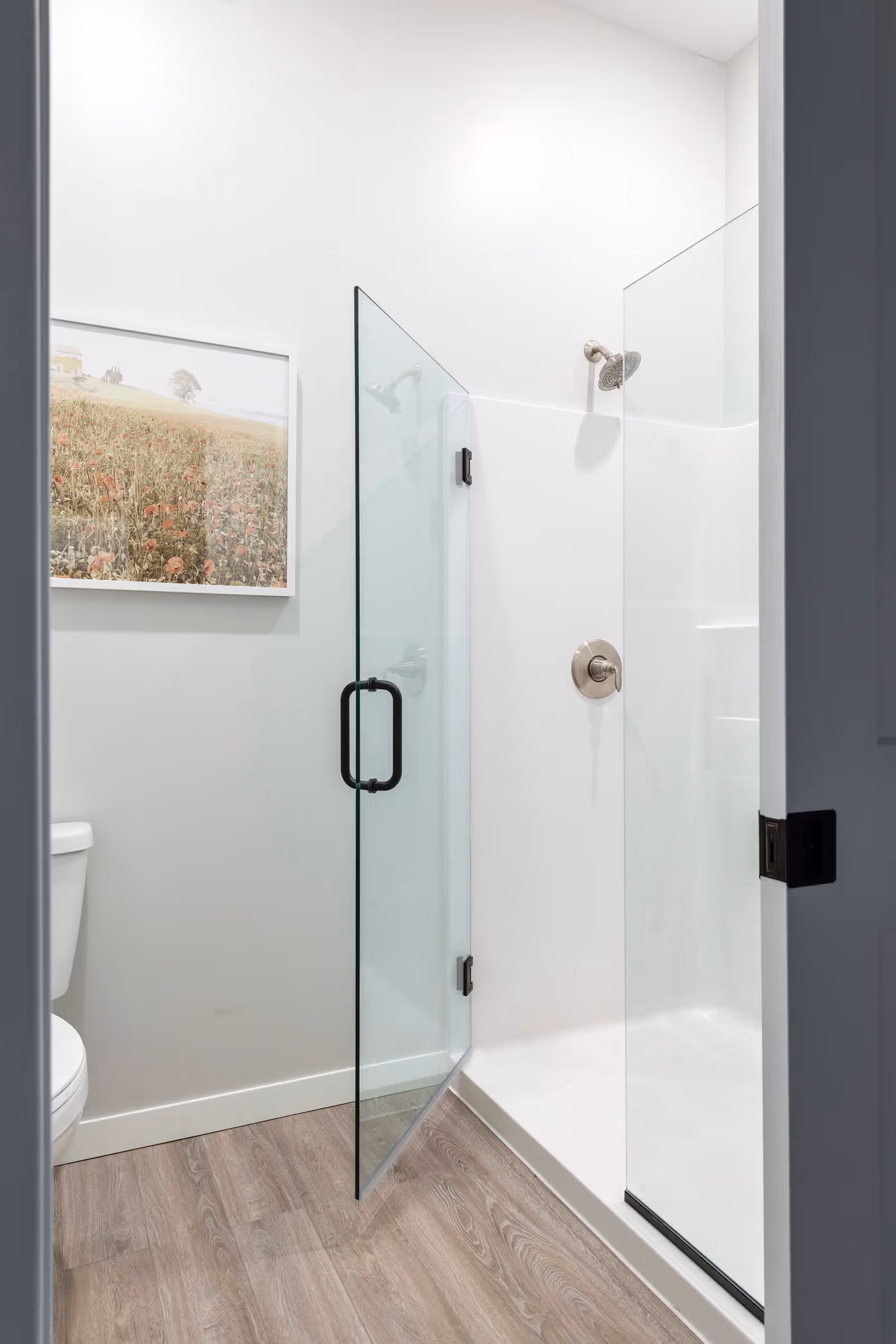 A modern bathroom with a glass shower enclosure featuring a metal handle and a silver showerhead. The floor has wood-like tiles, and a framed picture of a field with flowers hangs on the wall. A white toilet is partially visible on the left side.