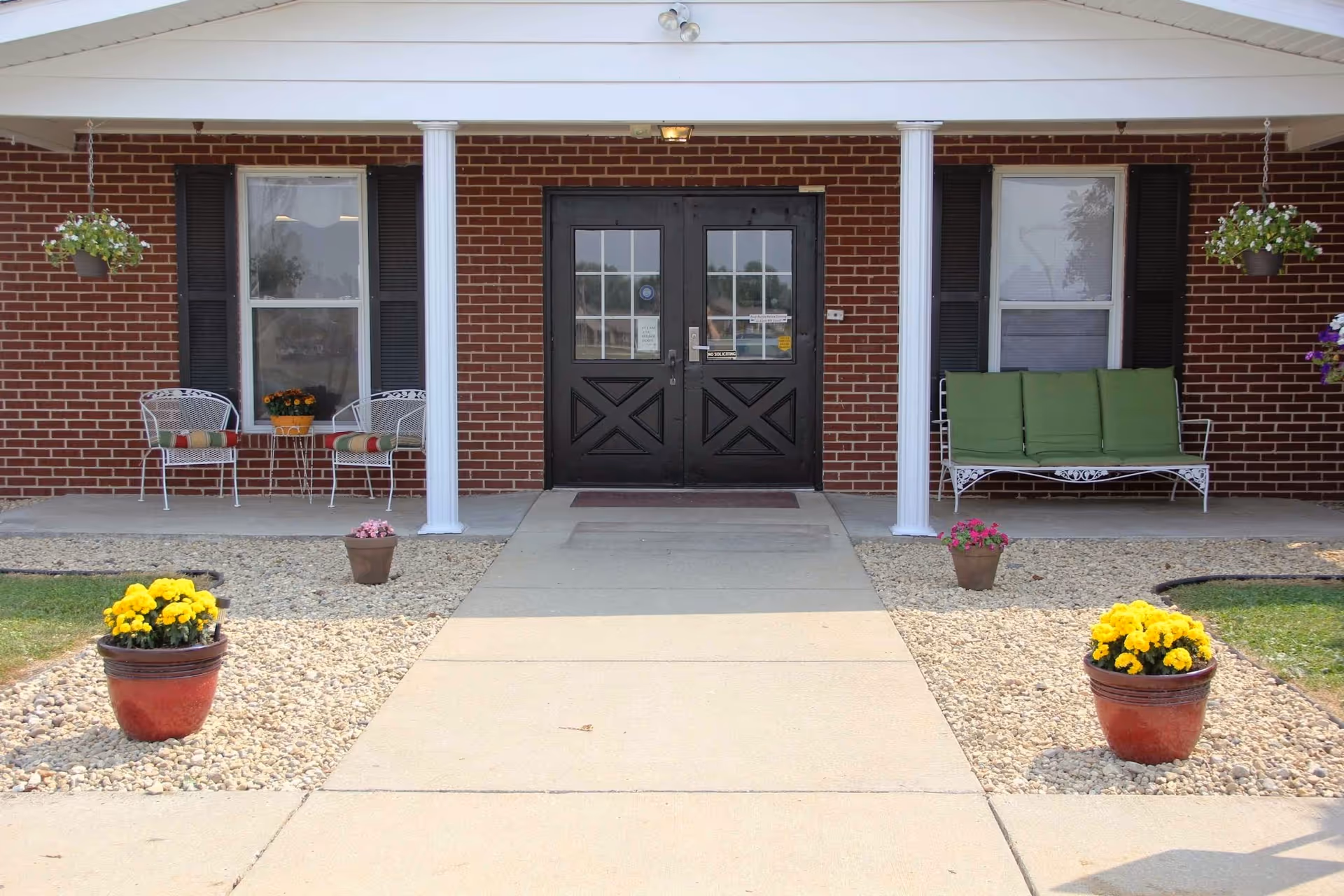 Brick building front with double entrance doors, a covered porch with white columns, chairs and a bench, and potted flowers along the walkway.