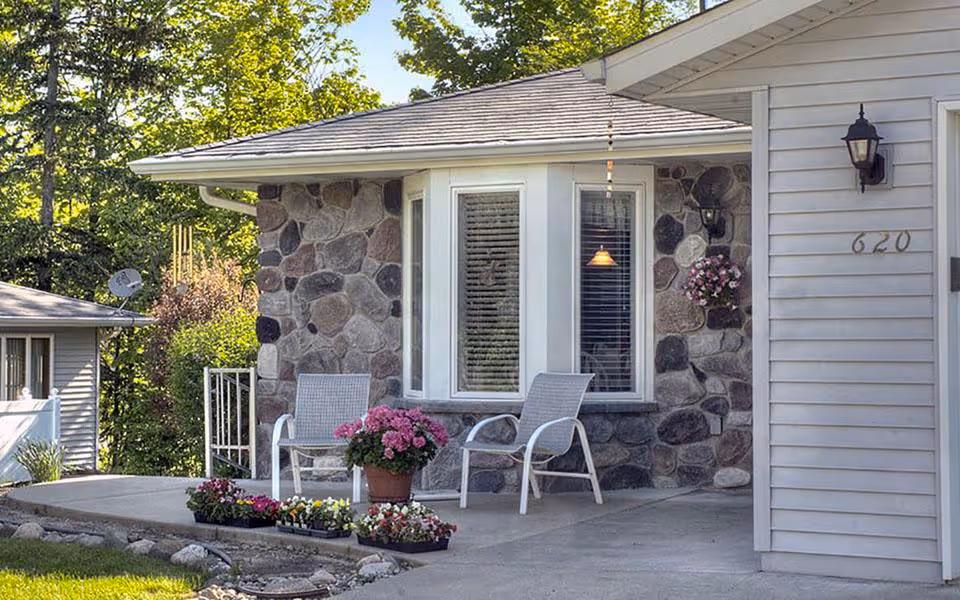 Outdoor patio area of a retirement community with two white chairs, a potted pink flower, and flower beds in front of a stone wall with a bay window. The house number 620 is visible on the white siding next to a black outdoor lantern. Trees and greenery are in the background.