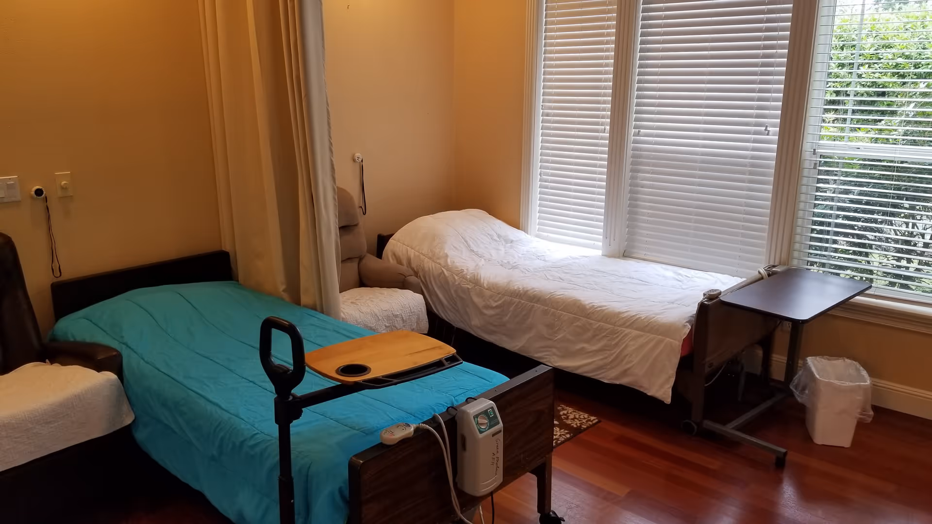A room with two hospital-style beds, one with a blue blanket and the other with a white blanket, positioned near large windows with white blinds. There is a small overbed table on the blue bed and another table near the white bed. The room has wooden flooring and beige walls, with a chair partially visible on the left side.