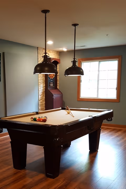 A room with a pool table set up for a game, featuring a rack of billiard balls and a cue stick on the table. Two black pendant lights hang from the ceiling above the table. There is a window with wooden trim letting in natural light, and a dartboard cabinet is mounted on a stone accent wall in the corner. The floor is wooden.