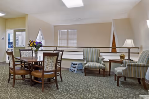 A sitting area in a senior living facility with a round wooden table surrounded by four chairs, a vase with flowers on the table, two striped armchairs, a side table with a lamp, and a carpeted floor. There is a railing behind the chairs and a window with blinds in the background.