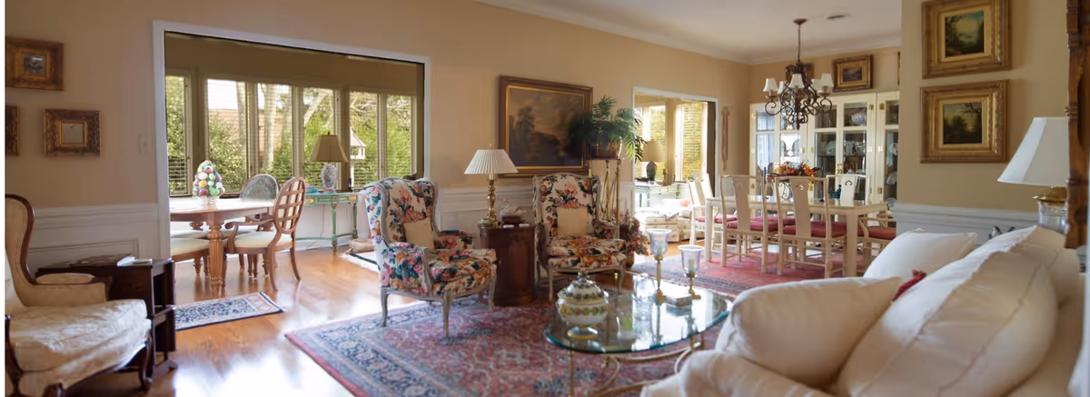 A bright and elegant living room and dining area with floral upholstered armchairs, a glass coffee table, a white sofa, wooden dining table with chairs, large windows letting in natural light, framed paintings on beige walls, and hardwood floors with area rugs.