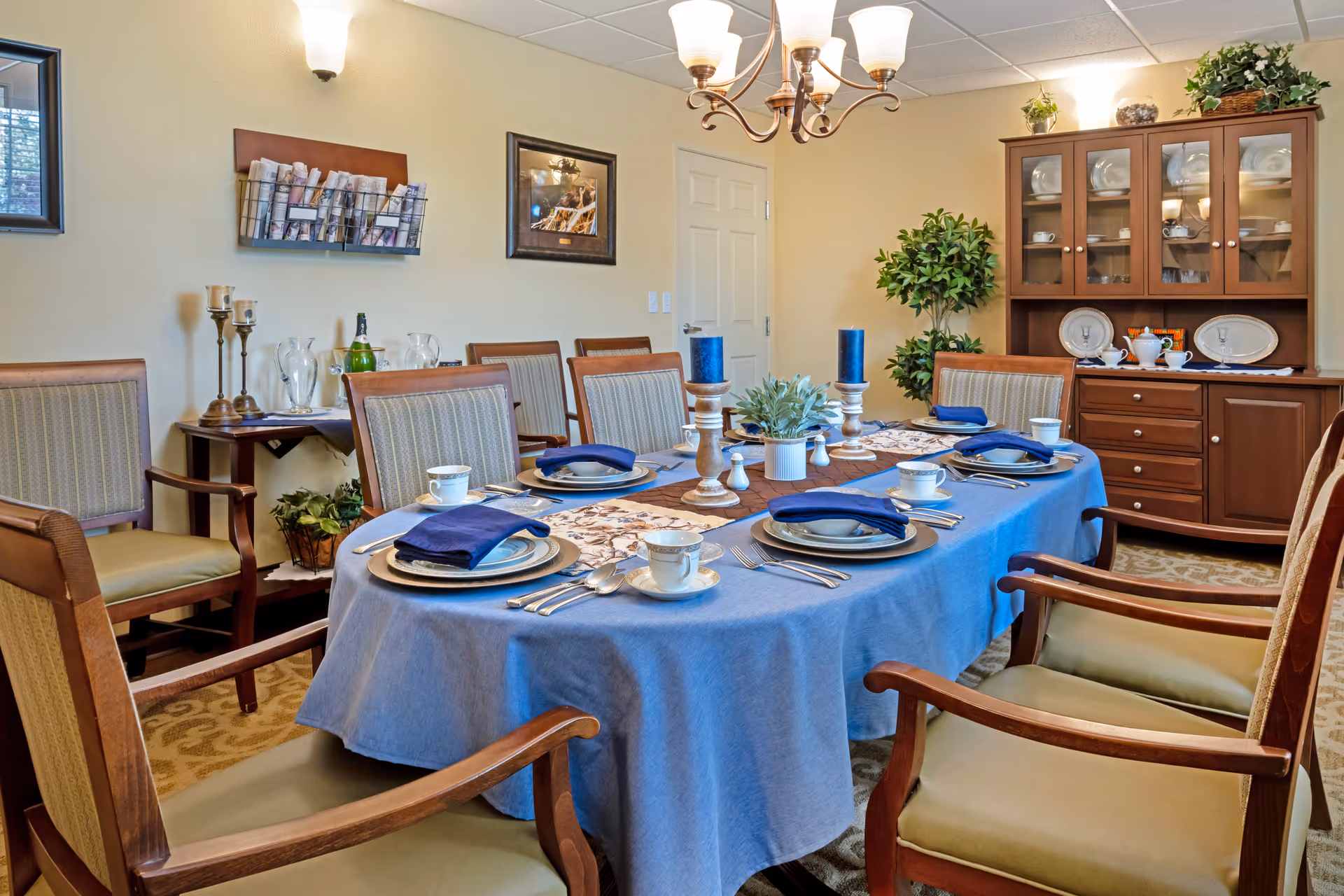 A dining room with a large oval table covered with a blue tablecloth and a decorative runner. The table is set with plates, cups, silverware, and blue napkins. There are wooden chairs with cushioned seats around the table. In the background, there is a wooden cabinet with glass doors displaying plates and cups, a potted plant, a wall-mounted magazine rack, and framed artwork on the wall. The room is softly lit by a chandelier and wall sconces.