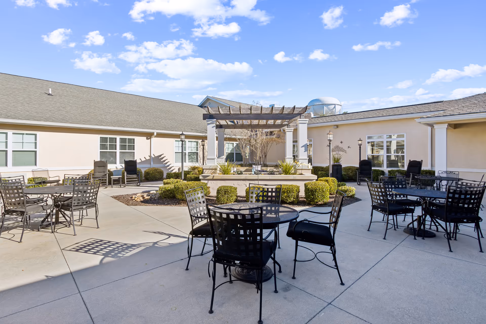 Outdoor courtyard area at Barclay House of Augusta featuring several black metal tables and chairs on a concrete patio. There is a central pergola structure surrounded by neatly trimmed bushes and plants. The building walls are light-colored with multiple windows, and the sky is clear with scattered clouds.