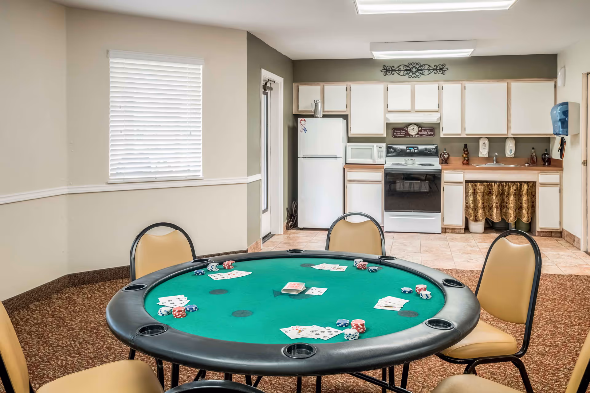 A room with a green poker table set with playing cards and poker chips, surrounded by beige chairs. In the background, there is a small kitchen area with white cabinets, a refrigerator, microwave, stove, and a sink with a decorative curtain underneath. The walls are painted in neutral tones and there is a window with closed blinds.