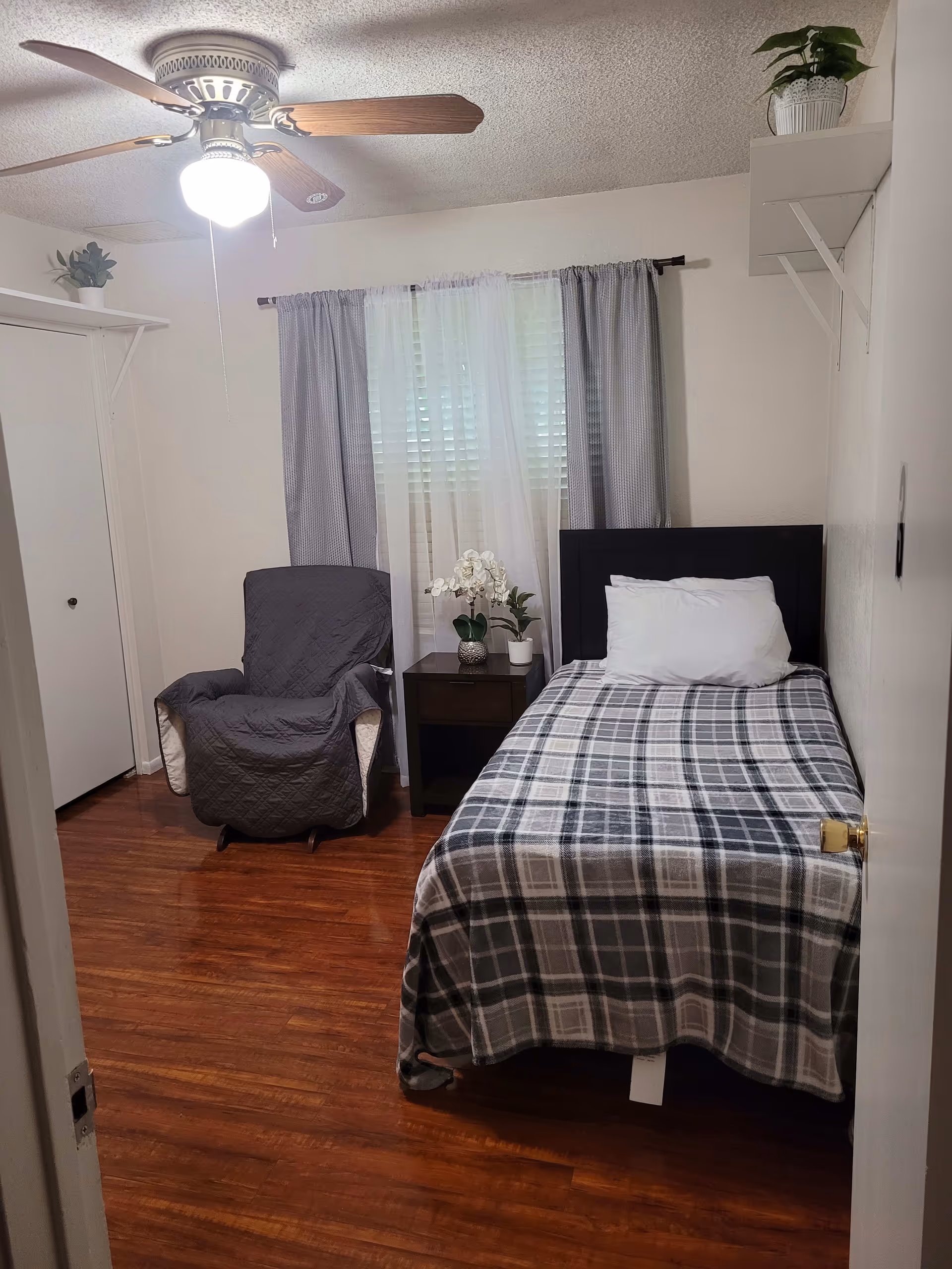 A small bedroom with a single bed covered in a gray and white plaid blanket and a white pillow. Next to the bed is a dark wooden nightstand with two potted plants and a white orchid. A gray armchair with a cover is positioned near the window, which has white blinds and sheer white curtains with gray drapes. The room has wooden flooring, white walls, a ceiling fan with a light, and two small shelves with potted plants on the walls.