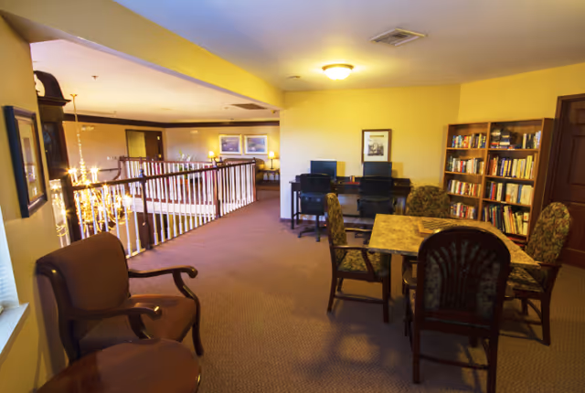 Interior view of a senior living facility common area with a table and four chairs, a bookshelf filled with books, two computer desks with chairs, and a railing overlooking a lower level. The walls are painted yellow and the carpet is brown.