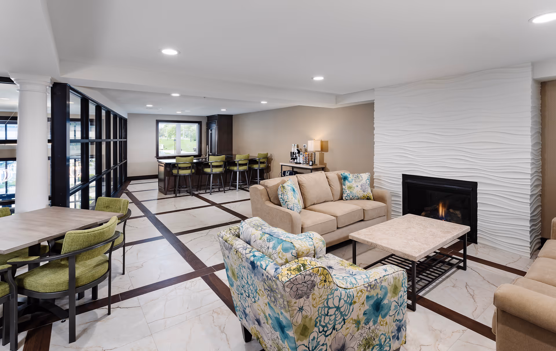 A modern senior community common area with beige sofas and a floral armchair arranged around a rectangular coffee table in front of a white textured fireplace. To the left, there is a dining table with green cushioned chairs and a bar area with high stools near a window. The floor has a white and dark brown tile pattern, and the space is well-lit with recessed ceiling lights.