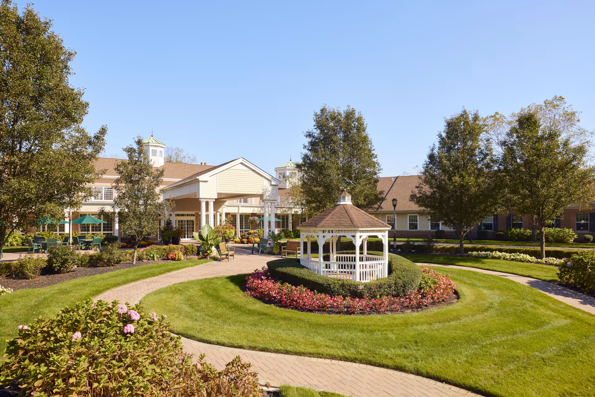 A well-maintained outdoor garden area at Brandywine Colts Neck by Monarch featuring a white gazebo surrounded by neatly trimmed hedges and colorful flowers. There are paved walkways curving through green lawns and trees, with a building in the background under a clear blue sky.