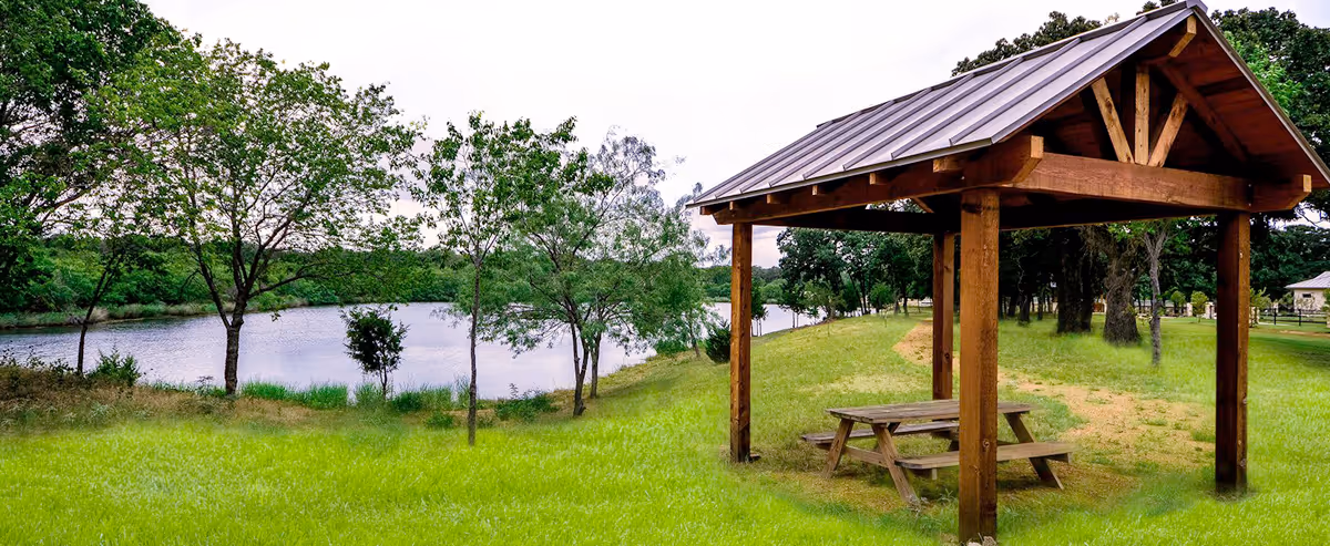 A wooden picnic shelter with a metal roof stands on a grassy area near a calm lake, surrounded by trees and greenery under an overcast sky.