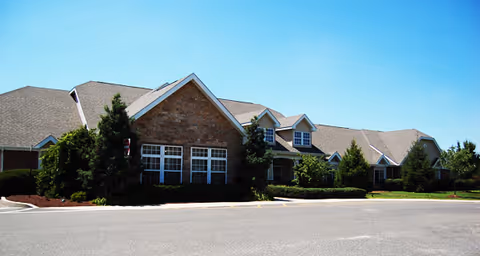 Brick single-story senior living building with gabled roofs, multiple windows, landscaped shrubs, and a paved driveway under a clear blue sky.