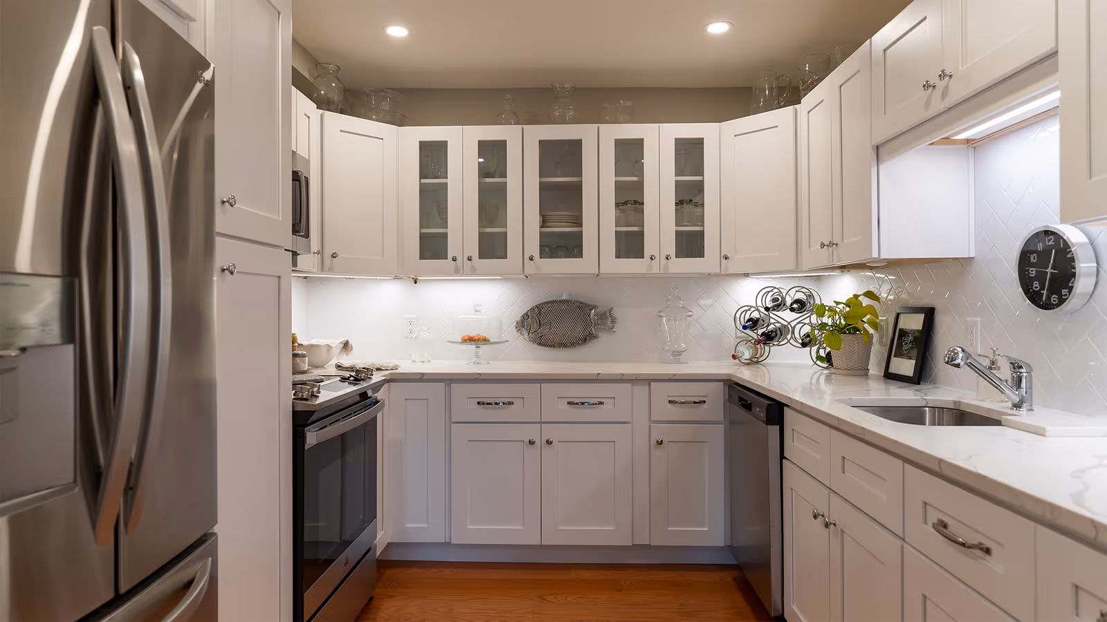 Modern kitchen with white cabinets, stainless steel refrigerator, oven, and dishwasher. The countertop is white with a built-in sink, a small plant, a wine rack, and a wall clock. The backsplash features white tiles arranged in a herringbone pattern.