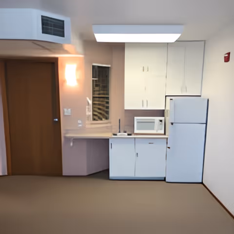 Small kitchenette with white cabinets, a refrigerator, microwave, sink and a closed wooden door to the left.
