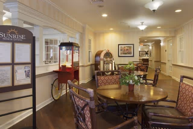 A cozy common area in a senior living facility featuring round wooden tables with cushioned chairs, a popcorn machine, a vintage jukebox, and framed artwork on the walls. The space has warm lighting and a hallway leading further into the building.